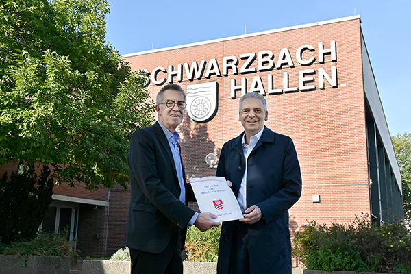 zwei Männer in Anzügen vor den Schwarzbachhallen halten eine Urkunde in den Händen