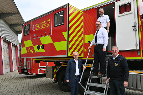 ein Feuerwehrauto an dessen Treppe vier Personen stehen