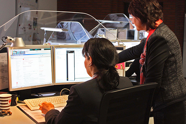 eine Frau mit einem Headset an einem Computer, neben ihr schaut ihr eine andere Frau bei der Arbeit zu. 
