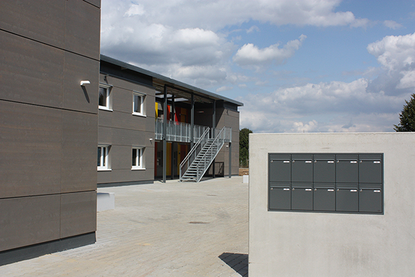 ein graues zweistöckiges Containergebäude. Im Vordergrund steht eine Mauer mit Briefkästen.