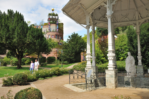 einen Park mit einer Mineralquelle im Vordergrund, während im Hintergrund ein buntes Haus zu sehen ist.