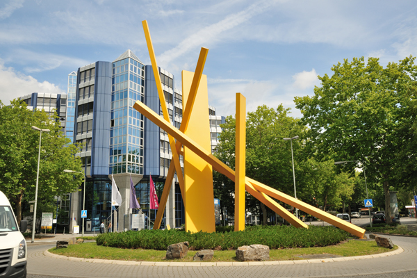 einen Kreisel, in dessen Mitte ein Kunstwerk aus gelben Stangen steht; im Hintergrund ist ein Büro-Hochhaus zu sehen.