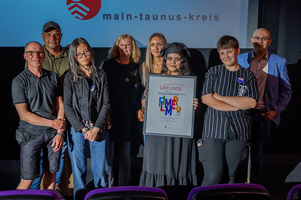 eine Gruppe Erwachsener und Jugendliche vor einer Kinoleinwand, die Jugendlichen halten eine Flimmer Urkunde