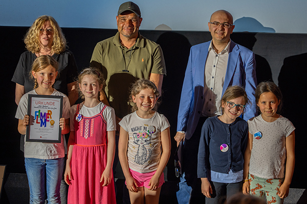 eine Gruppe Erwachsener und Kinder vor einer Kinoleinwand, die Kinder halten eine Flimmer Urkunde