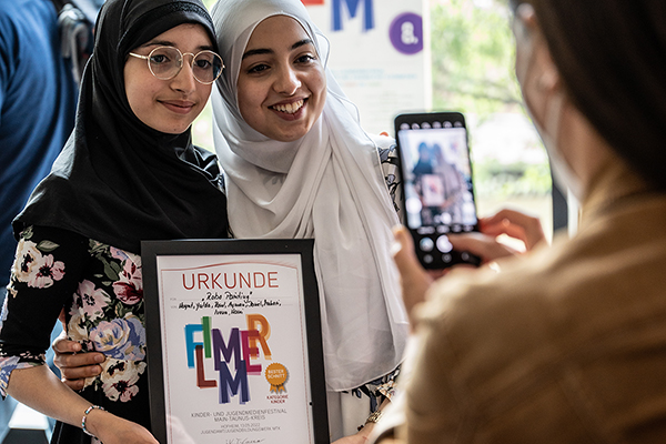 zei Mädchen mit Kopftuch halten eine Flimmer Urkunde, eine Frau macht ein Foto der Beiden mit dem Handy