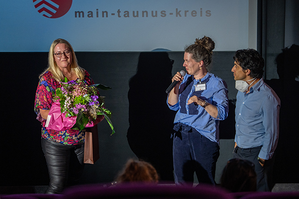 zwei Frauen und ein Mann vor einer Leinwand, eine hält ein Mikrofon, die andere einen Blumenstrauß