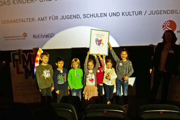 eine Gruppe Kinder auf einer Bühne, die eine große Urkunde in die Luft halten