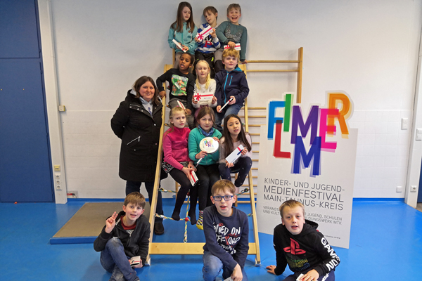 eine Gruppe Kinder, die in einem Turnraum auf einem Klettergerüst sitzen; daneben eine Frau und ein Plakat mit dem Text FLIMMER