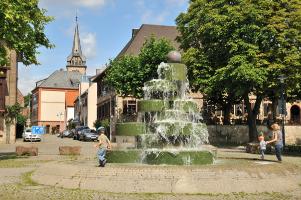 einen Brunnen aus Stein, an dem Kinder spielen; im Hintergrund liegt eine Altstadt, aus der ein Kirchturm herausragt.