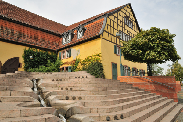 eine breite Steintreppe, in deren Mitte ein Wasserlauf nach unten fließt; im Hintergrund steht ein großes, gelbes Fachwerkhaus.