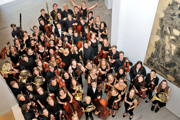 ein Orchester bestehend aus jungen Musikern, die mit ihren Instrumente in einem Raum stehen und nach oben in die Kamera schauen