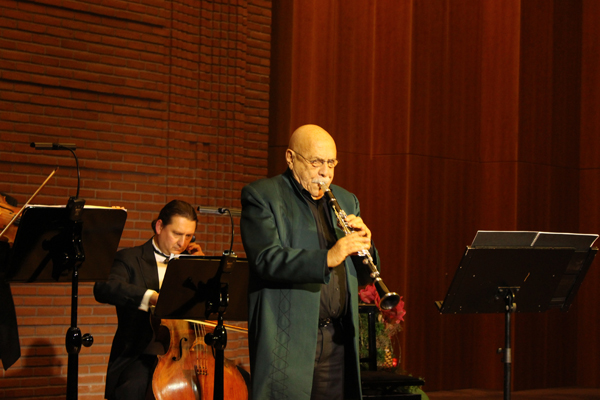 ein Mann mit einer Klarinette; im Hintergrund Notenständer und Mitglieder eines Orchesters