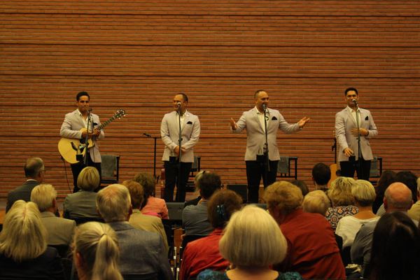 vier Männer vor Mikrofonen, einer mit Gitarre, auf einer Bühne vor Publikum
