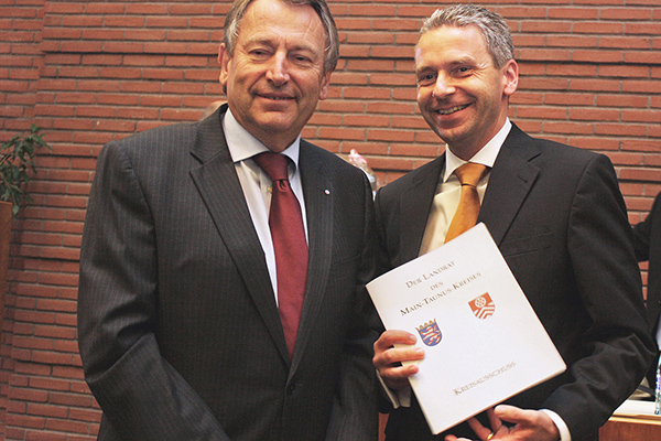 zwei Männer in einem Saal; der rechte hält eine Urkunde mit dem Wappen des Main-Taunus-Kreises in der Hand.