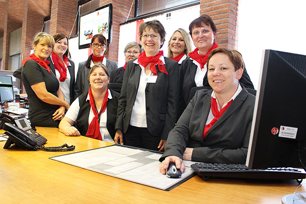 neun Frauen an einem Empfangstisch neben einem Computermonitor. Sie tragen dunkle Jacken, weiße Blusen und rote Halstücher.