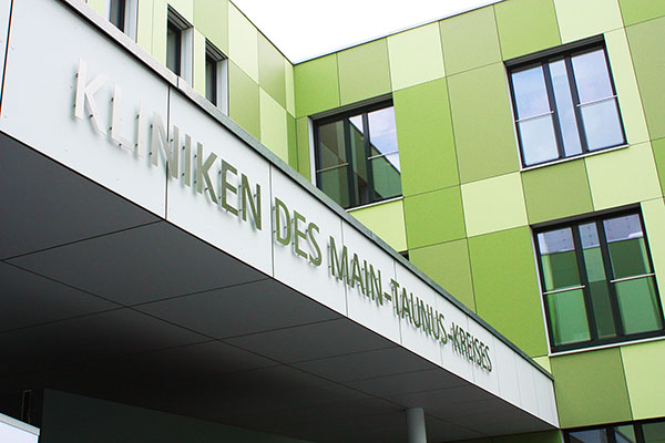 ein grünes mehrstöckiges Gebäude. Über dem Eingangsportal prangt die Schrift Kliniken des Main-Taunus-Kreises.