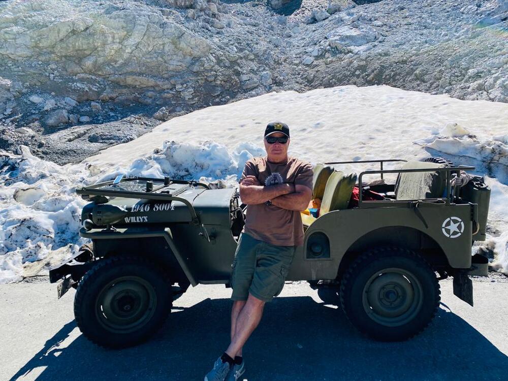 Olivgrüner, militärischer Geländewagen steht vor einem Eisglätscher in den Bergen, davor steht ein Mann in kurzen Hosen