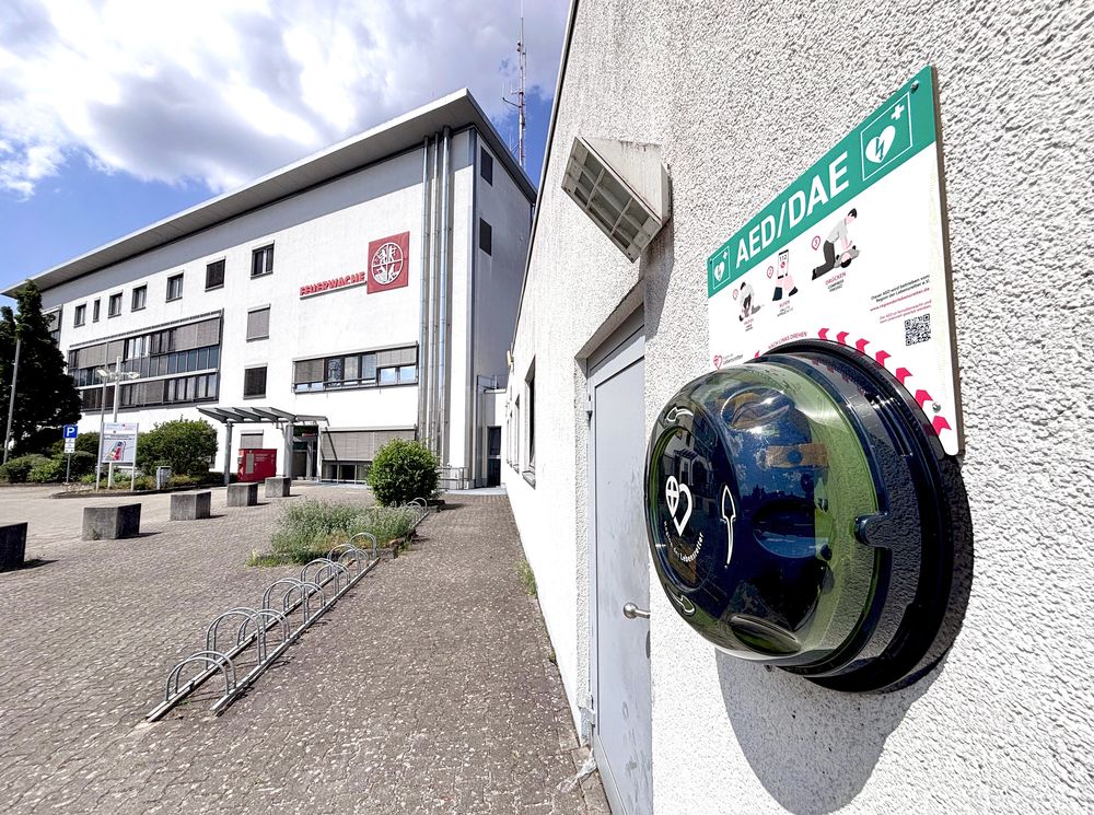 Ein AED befestigt an einer Hauswand; im Hintergrund die Feuerwehrwache in Hofheim