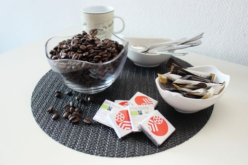ein Tisch mit einer Schale Kaffeebohnen, einigen Tafeln Schokolade, einer Schale Zucker, einer Tasse und einigen Löffeln
