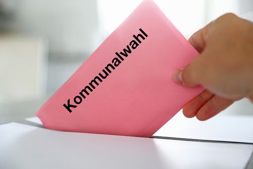 Eine Hand, die einen rosafarbenen Umschlag mit dem Text Kommunalwahl in eine Wahlurne steckt