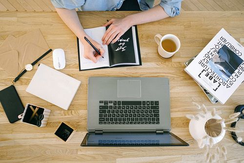 ein Arbeitstisch mit Laptop, Tee und weiteren Büroutensilien, jemand schreibt in ein Kalenderbuch
