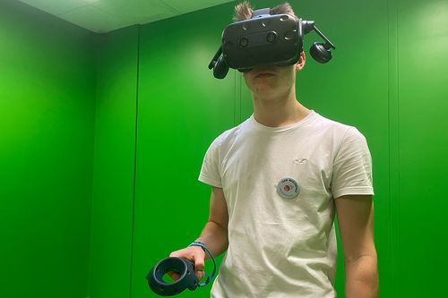 einen jungen Mann mit VR-Brille und Controller in einem grünen Raum
