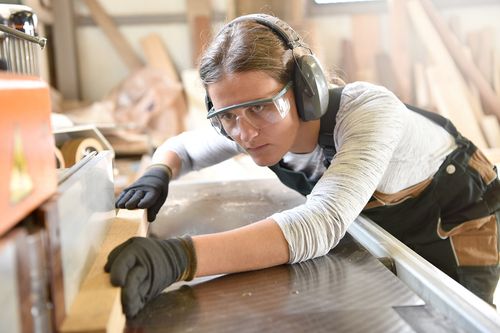 Zu sehen ist eine junge Frau, die mit Augen- und Ohrenschutz auf dem Kopf ein Stück Holz zurechtsägt.
