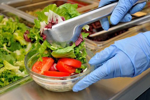 Ein Foto einer Schul-Essensausgabe: Zu sehen ist eine Salatschüssel, die gerade mit Salatblättern und Tomaten durch das Catering-Personal gefüllt wird
