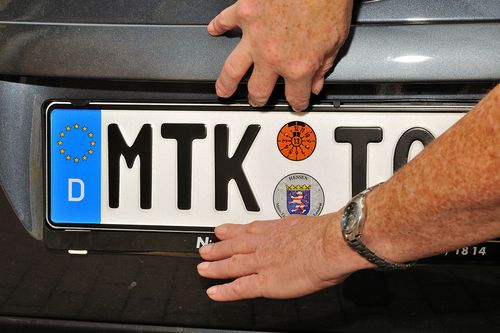 Ein Nahaufnahme eines KFZ-Kennzeichens, das gerade durch eine Hand am Auto angebracht wird
