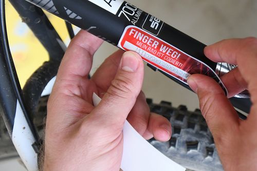 Eine Person bringt einen roten Sicherheitsaufkleber mit der Aufschrift „Finger weg! Mein Rad ist codiert“ an einem Fahrrad an
