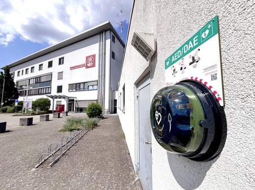 Ein AED befestigt an einer Hauswand; im Hintergrund die Feuerwehrwache in Hofheim