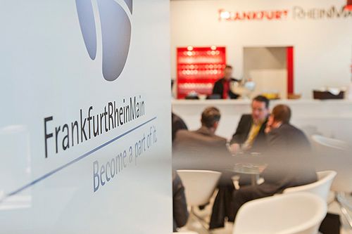 Im Vordergrund Logo FrankfurtRheinMain im Hintergrund Personen am Tisch und eine weitere am Empfang