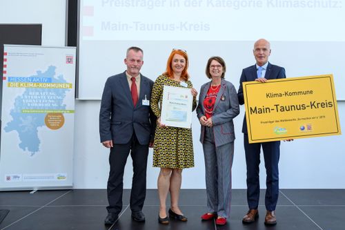 Zwei Frauen und zwei Männer, eine hält eine Urkunde in der Hand, ein zweiter ein großes Ortsschild mit dem Text Main-Taunus-Kreis