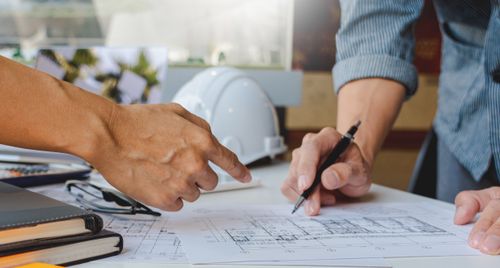 Zwei Personen arbeiten gemeinsam an Bauplänen auf einem Tisch; eine zeigt mit dem Finger auf Details