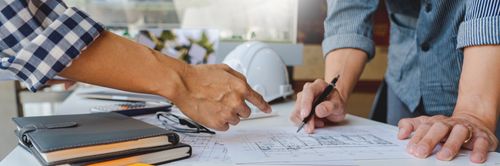Zwei Personen arbeiten gemeinsam an Bauplänen auf einem Tisch; eine zeigt mit dem Finger auf Details